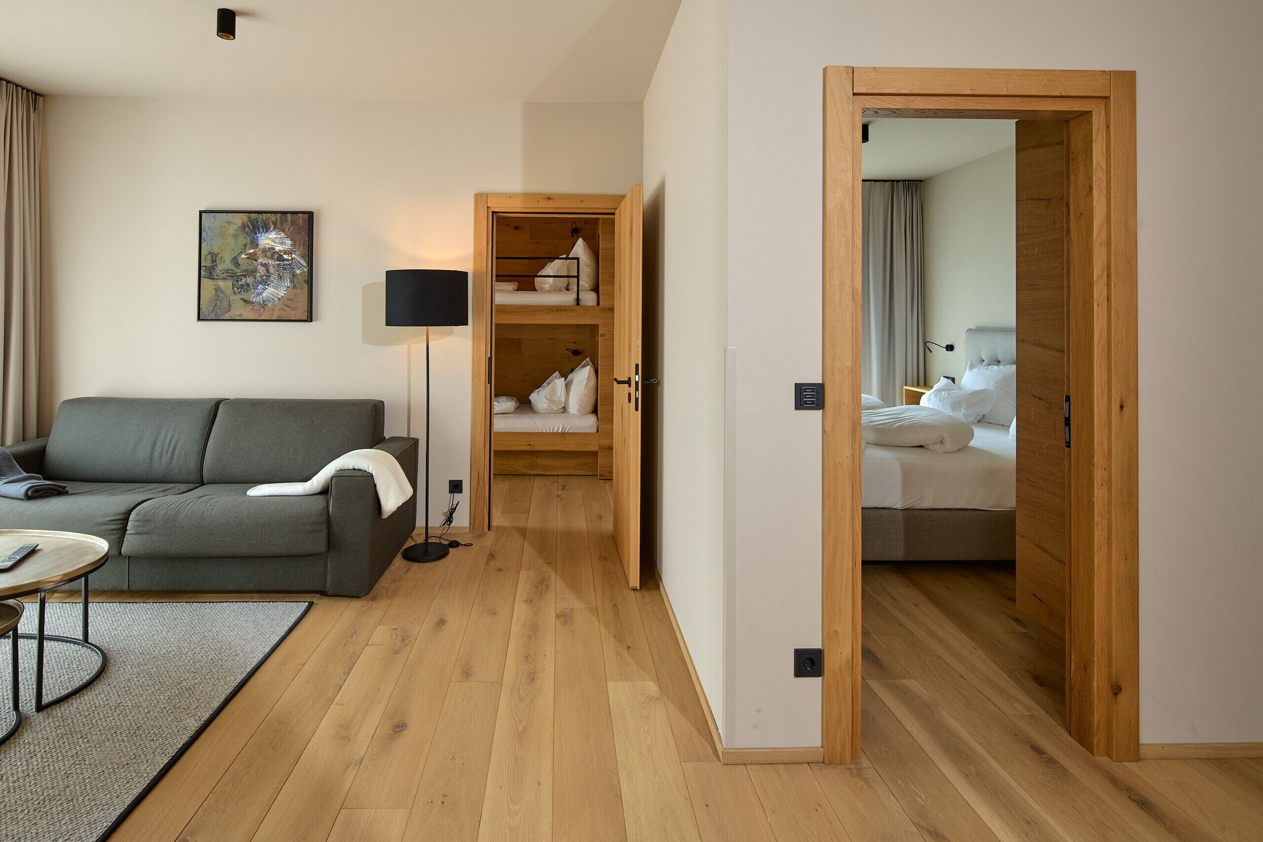 A modern hotel room with a grey sofa, a wooden floor, a painting, a lamp, and a wooden wardrobe with a bed.