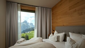 Ett hotellrum med en stor säng, ett träfärgat sänggavel och utsikt över bergen genom tunna, genomskinliga gardiner.