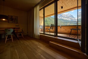A room with wooden flooring, a dining table with chairs, a large window with a view of mountains, and a bench by the window. - 8018 Summit Residence 18 (Schröcken)