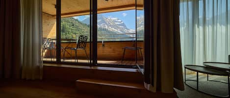 Ein Zimmer mit großem Fenster und Blick auf eine gebirgige Landschaft, Holzboden und einem kleinen Tisch mit Stühlen.