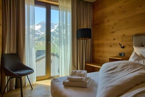 Habitació d'hotel amb un llit, una cadira, una taula de nit, una làmpada i vista a les muntanyes a través d'una finestra gran.