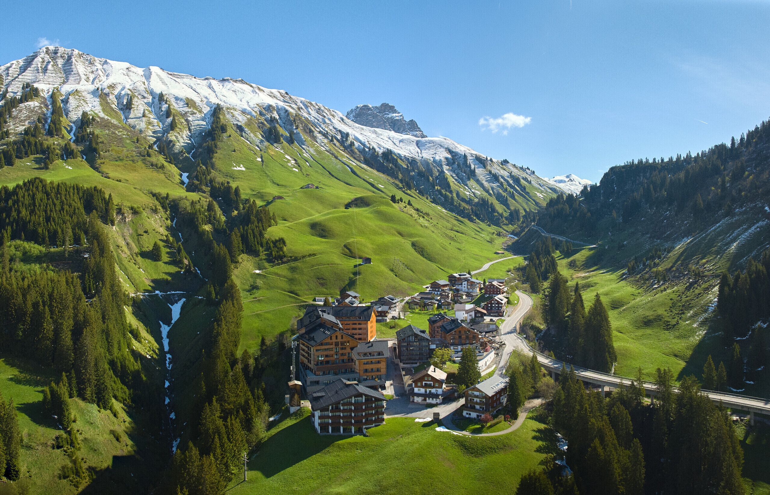 A mountainous village with a winding road, buildings with traditional architecture, and a valley with greenery and a river.