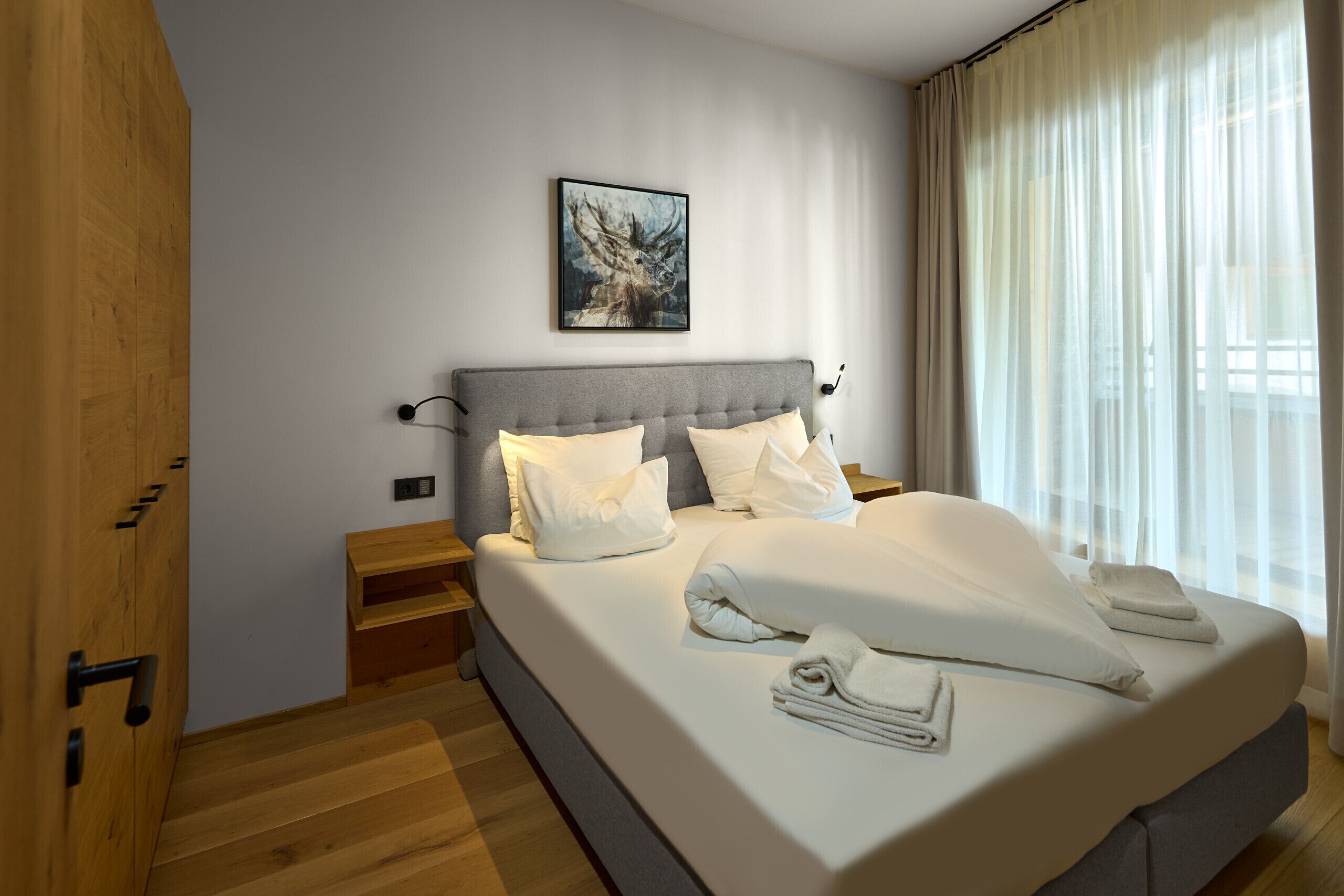 A modern bedroom with a large bed, a headboard, a bedside table, a nightstand, a painting on the wall, and a window with curtains.