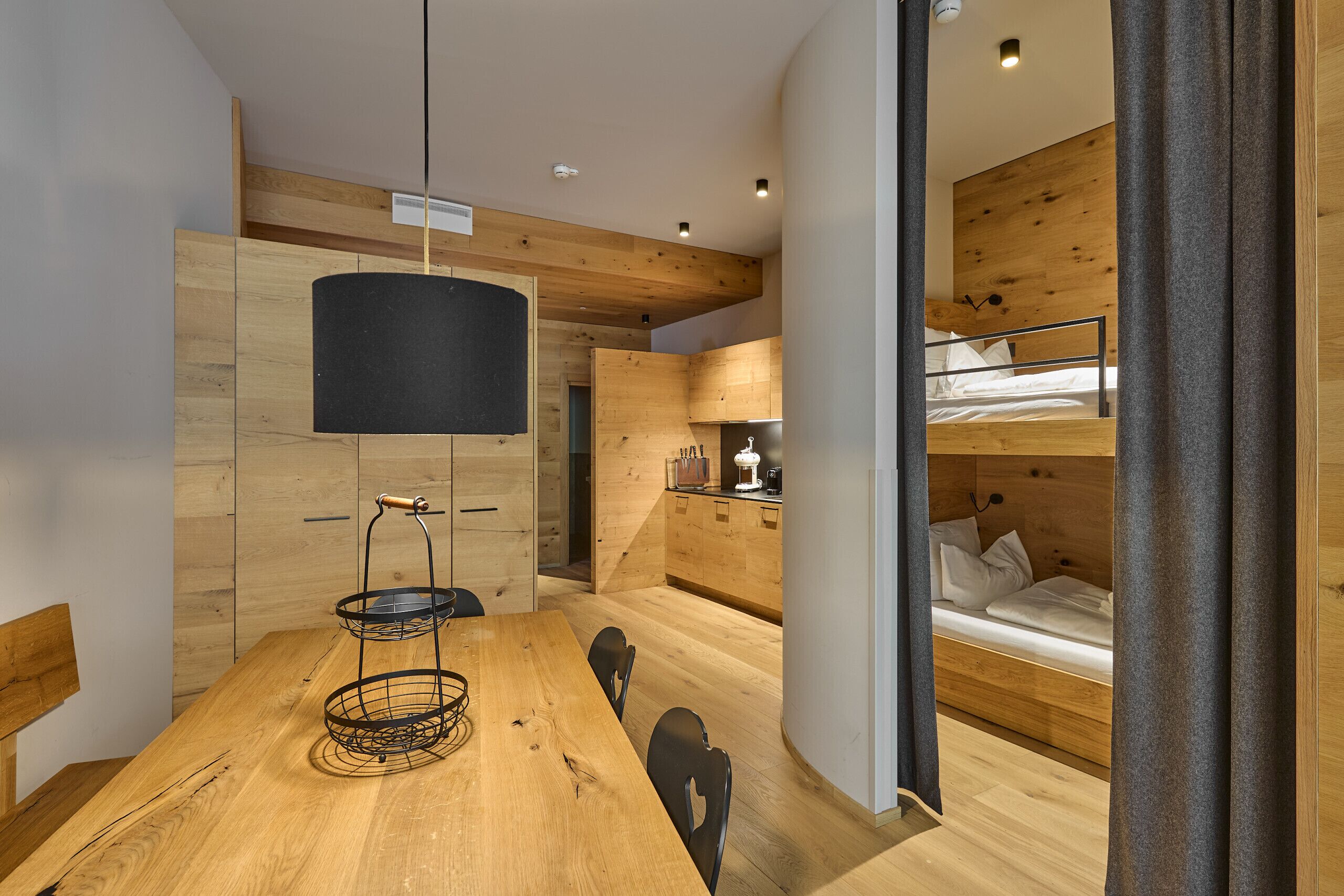Ein modernes Interieur mit einem Holzespeisetisch, einer schwarzen Hängeleuchte und einem Etagenbett mit Holz Kopfteil.
