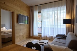 A modern hotel room with a wooden interior, a flat-screen TV mounted on the wall, a sofa, a small round table with a remote control, and a bed with a neatly folded towel. - 8023 Alpin Retreat Grand 23 (Schröcken)