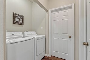A laundry room with two front-loading washers and dryers, a door with a brass doorknob, and a wall-mounted sign above the appliances. - The Gathering House On May (Fort Smith)