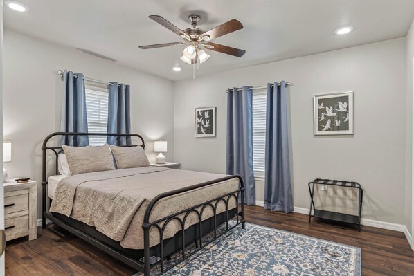 A bedroom with a bed, a nightstand, a bedside table, a ceiling fan, and a window with curtains. - The Gathering House On May (Fort Smith)