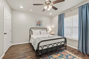 A bedroom with a bed, a ceiling fan, a window with curtains, and a wall-mounted light. - The Gathering House On May (Fort Smith)
