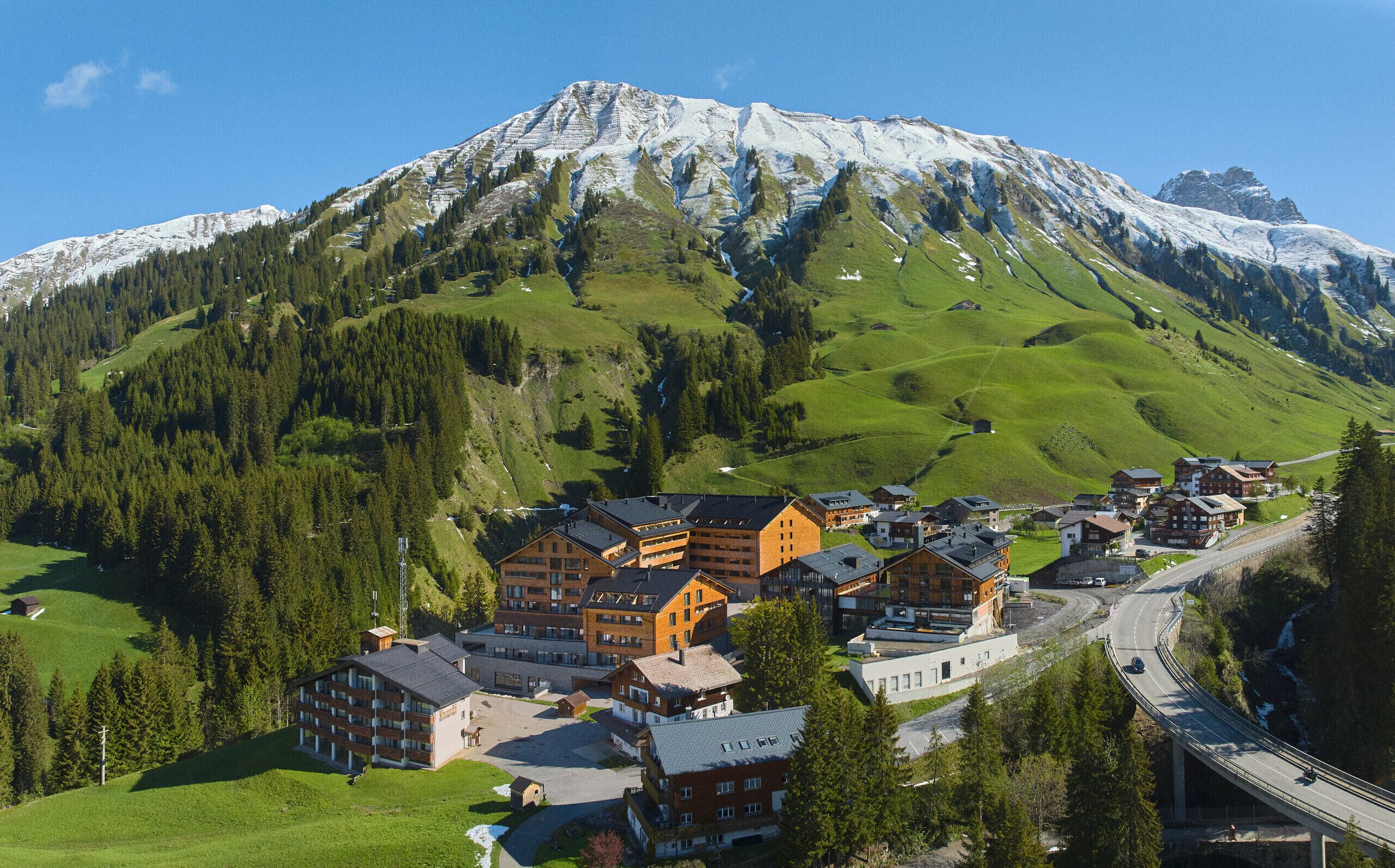 Une ville de montagne où se côtoient des bâtiments traditionnels et modernes, avec une route sinueuse et, en arrière-plan, un sommet enneigé qui domine le paysage.