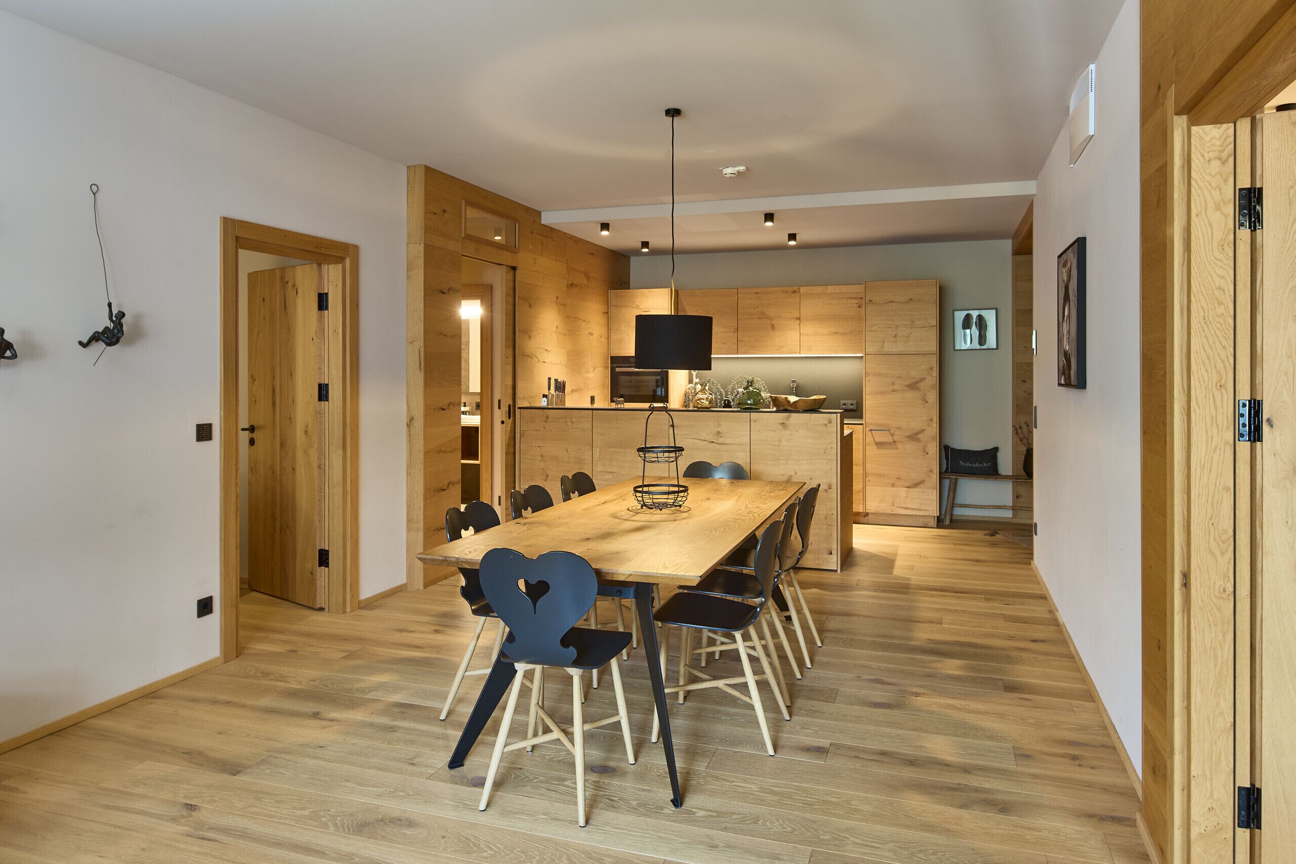 A modern kitchen with wooden cabinets and a central island, a dining area with a wooden table and chairs, and a hallway with wooden flooring.