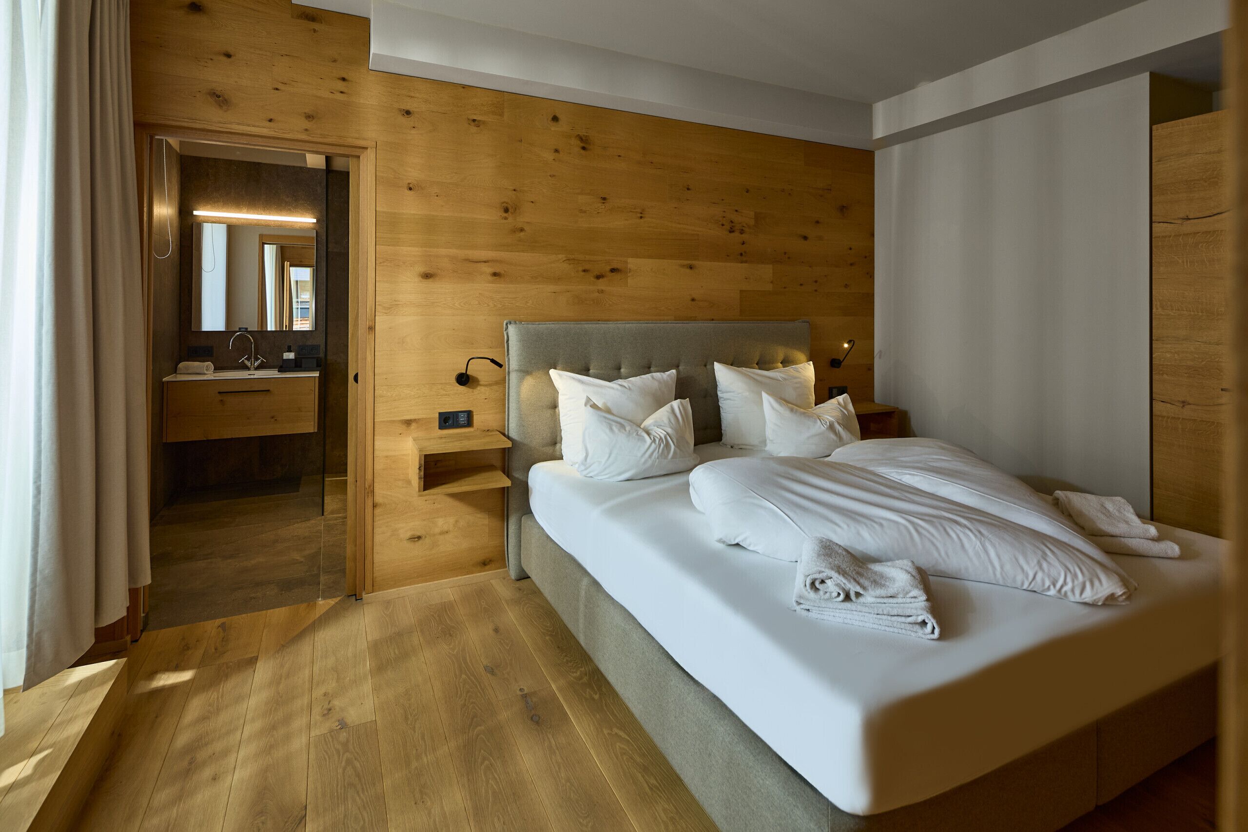 Een moderne slaapkamer met een houten hoofdbord, een groot bed met witte lakens, een nachtkastje en een spiegel boven de wastafel.