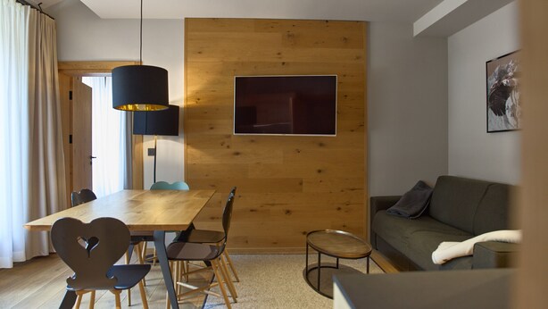 A modern living room with a wooden accent wall, a dining area with a wooden table and chairs, a sofa, and a mounted artwork. - 8031 Sky Residence 31 (Schröcken)