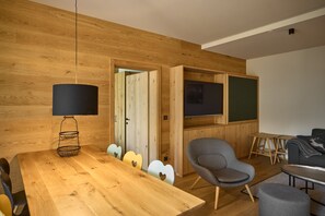 Ein modernes Wohnzimmer mit einer Holzwand, einem Esstisch mit Stühlen, einem grauen Sessel, einer kleinen Holz-TV-Konsole und einer Lampe.