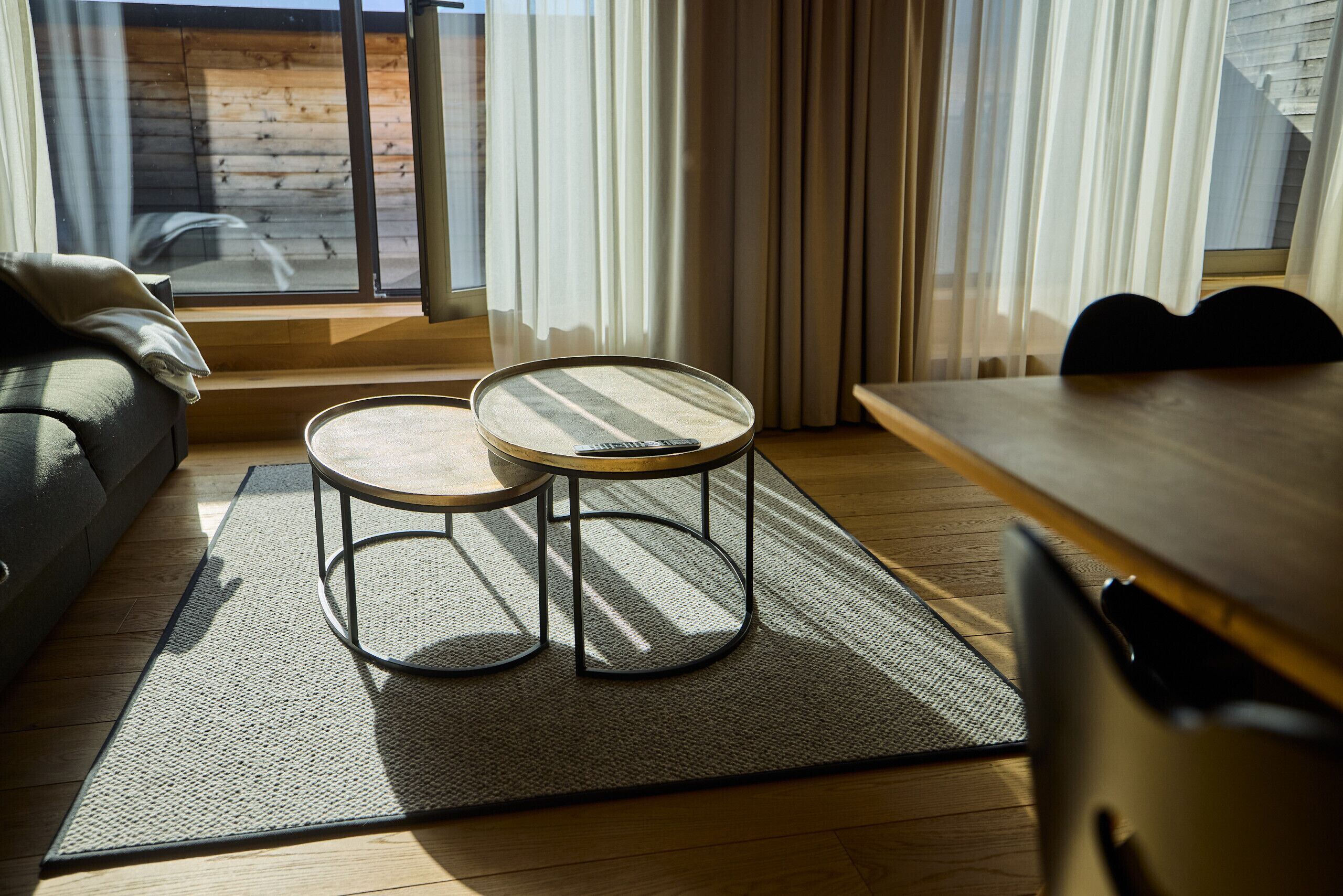 A modern living room with a sofa, two round tables, a wooden dining table, and a view through a large window.