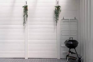 A white wooden wall with two hanging plants, a grill, and a window with a metal grill. - Linneby , fri Parkering (Uddevalla)