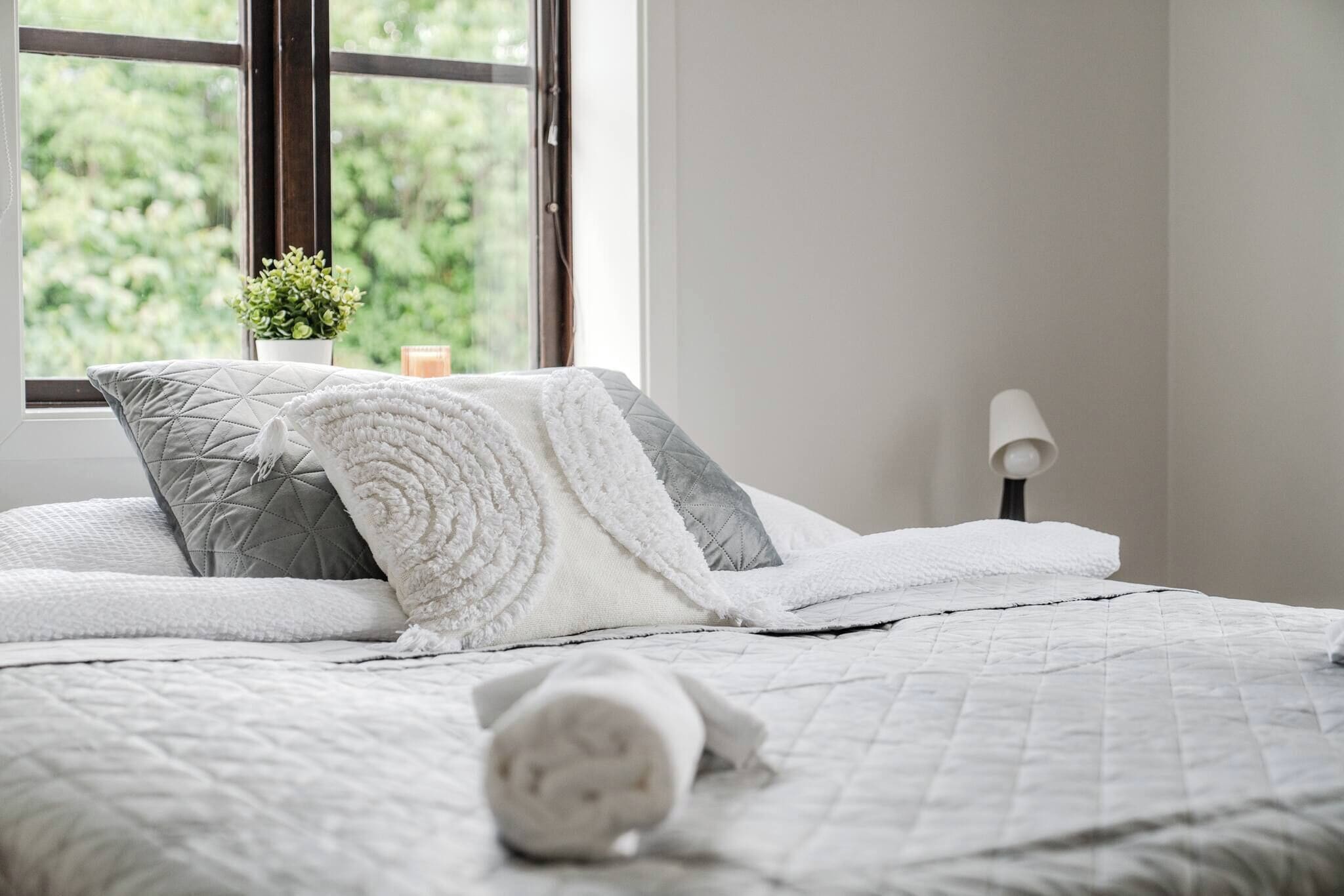 A neatly made bed with a quilted white bedspread and pillows, a bedside lamp, and a window with a view of greenery.