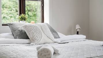 A neatly made bed with a quilted white bedspread and pillows, a bedside lamp, and a window with a view of greenery.