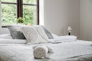 A neatly made bed with a quilted white bedspread and pillows, a bedside lamp, and a window with a view of greenery.