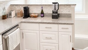 Eine Küche mit weißen Schränken, einer Granitarbeitsplatte, einer Kaffeemaschine, einem Mixer und einem Fenster mit Jalousien.