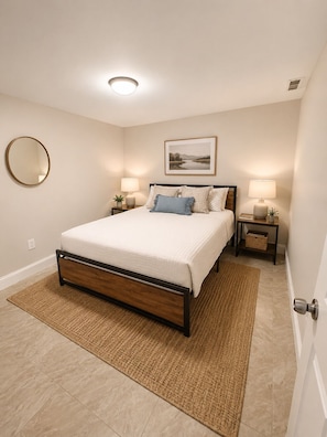 A bedroom with a bed, two bedside tables, a nightstand, a wall-mounted mirror, a framed picture, and a door. - Beautiful 3 bedroom house - Boston Beachside House Near Shore & Train (Revere)