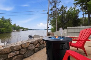 A lakeside outdoor seating area with a stone wall, a fire pit, and red chairs.