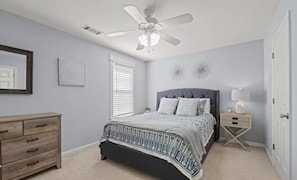 Ein Schlafzimmer mit einem Bett, einem Schminktisch, einem Nachttisch, einem Deckenventilator und einem Fenster mit Jalousien.
