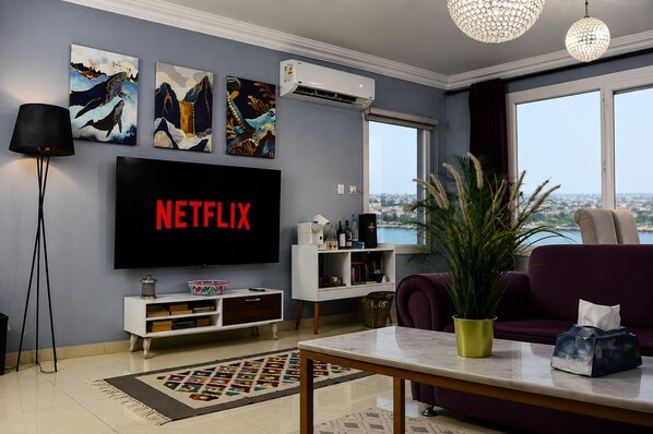 A living room with a flat-screen TV displaying 'NETFLIX,' a large window with a city view, and a potted plant.
