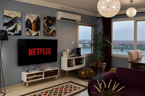 A living room with a flat-screen TV, artwork, a plant, and a view of a cityscape through a window.