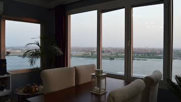 A room with a large window overlooking a river and cityscape, a sofa, a small table, and a potted plant.