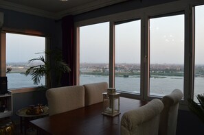 A room with a large window overlooking a river and cityscape, a sofa, a small table, and a potted plant.