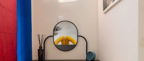 A room with a desk, two yellow chairs, a round mirror, a neon cloud light, and a framed picture on the wall.