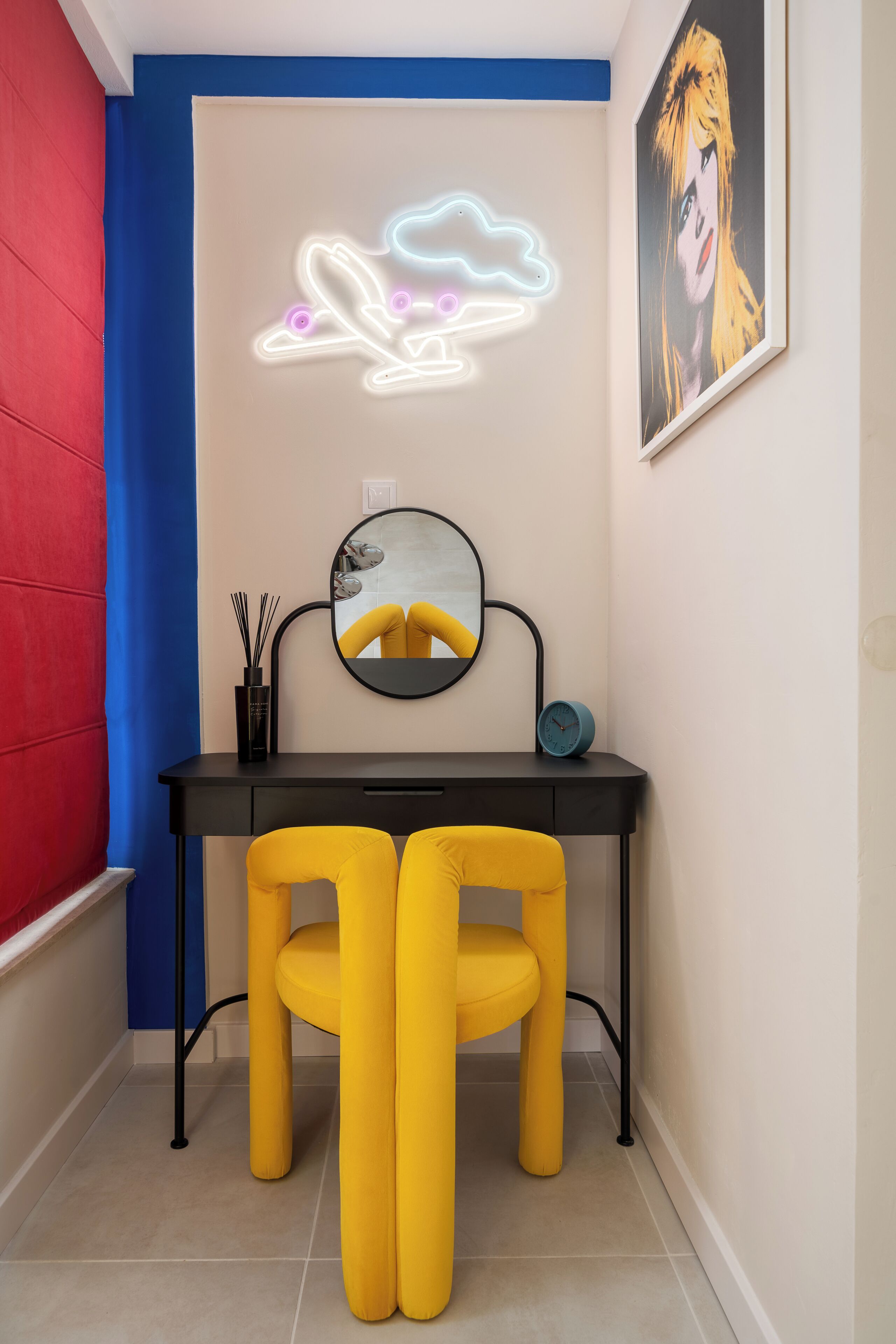 Una habitación con un escritorio, dos sillas amarillas, un espejo redondo, una luz de neón y un cuadro en la pared.