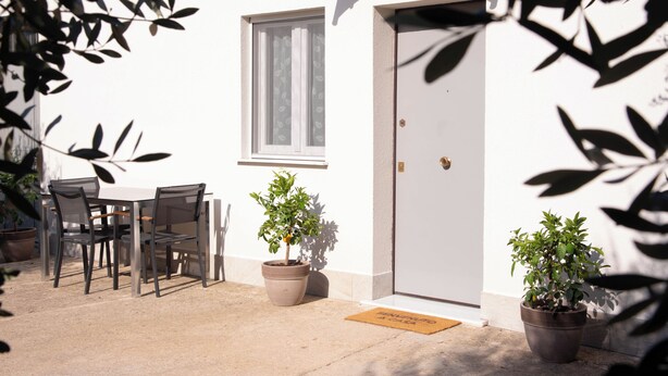 Área externa com mesas e cadeiras, dois vasos de plantas e uma porta com tapete.