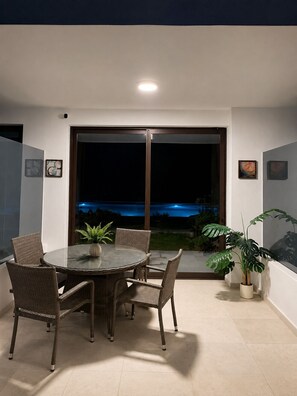 A dining area with a glass table and wicker chairs, a potted plant, and a view of a body of water at night. - Luxury 1-Br Condo at Hard Rock Punta Cana — Resort Living, 15 mins from Downtown (Punta Cana)