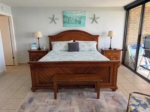 Quarto com cama de madeira, duas mesas de cabeceira com luminárias, um banco no pé da cama, pintura de uma cena de praia e decorações de estrelas-do-mar na parede.