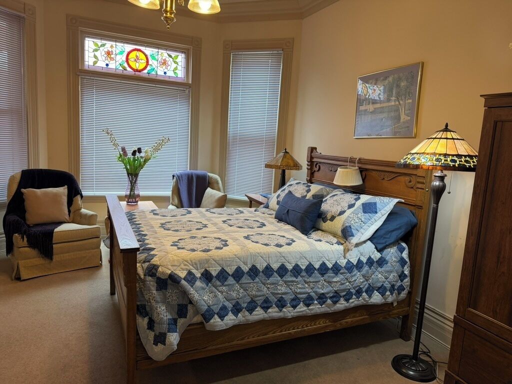 A bedroom with a bed, two chairs, a lamp, and a window with a stained glass design.