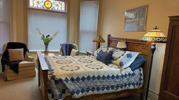A bedroom with a bed, two chairs, a lamp, and a window with a stained glass design.