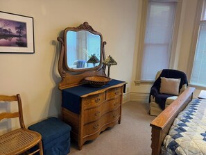 Une chambre à coucher avec une commode en bois, un miroir, une chaise, un lit recouvert d’une courtepointe et une fenêtre avec des stores.