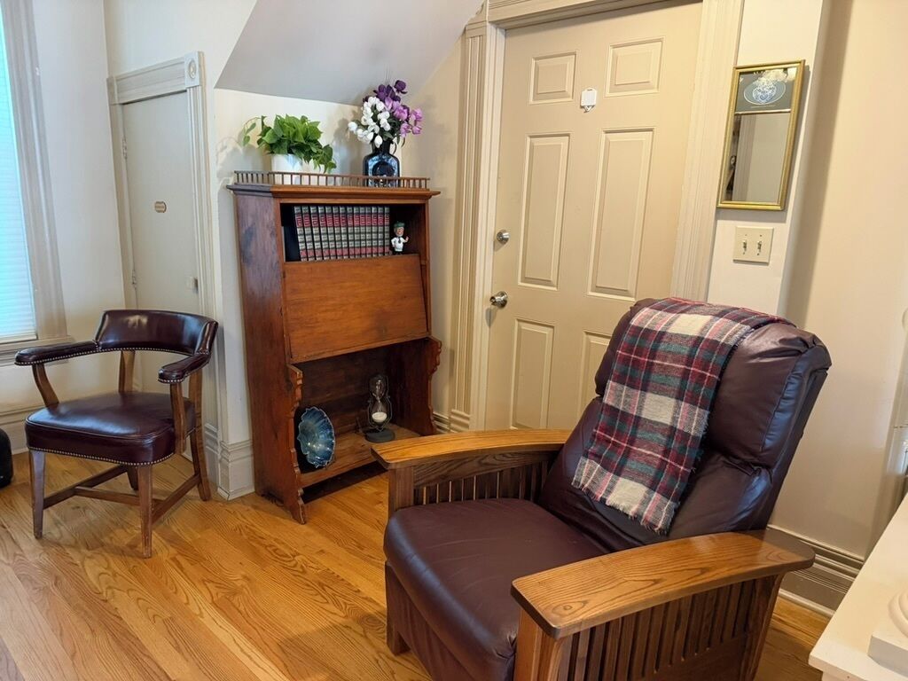 A room with a wooden bookshelf, a leather armchair with a plaid throw, and a wooden chair.