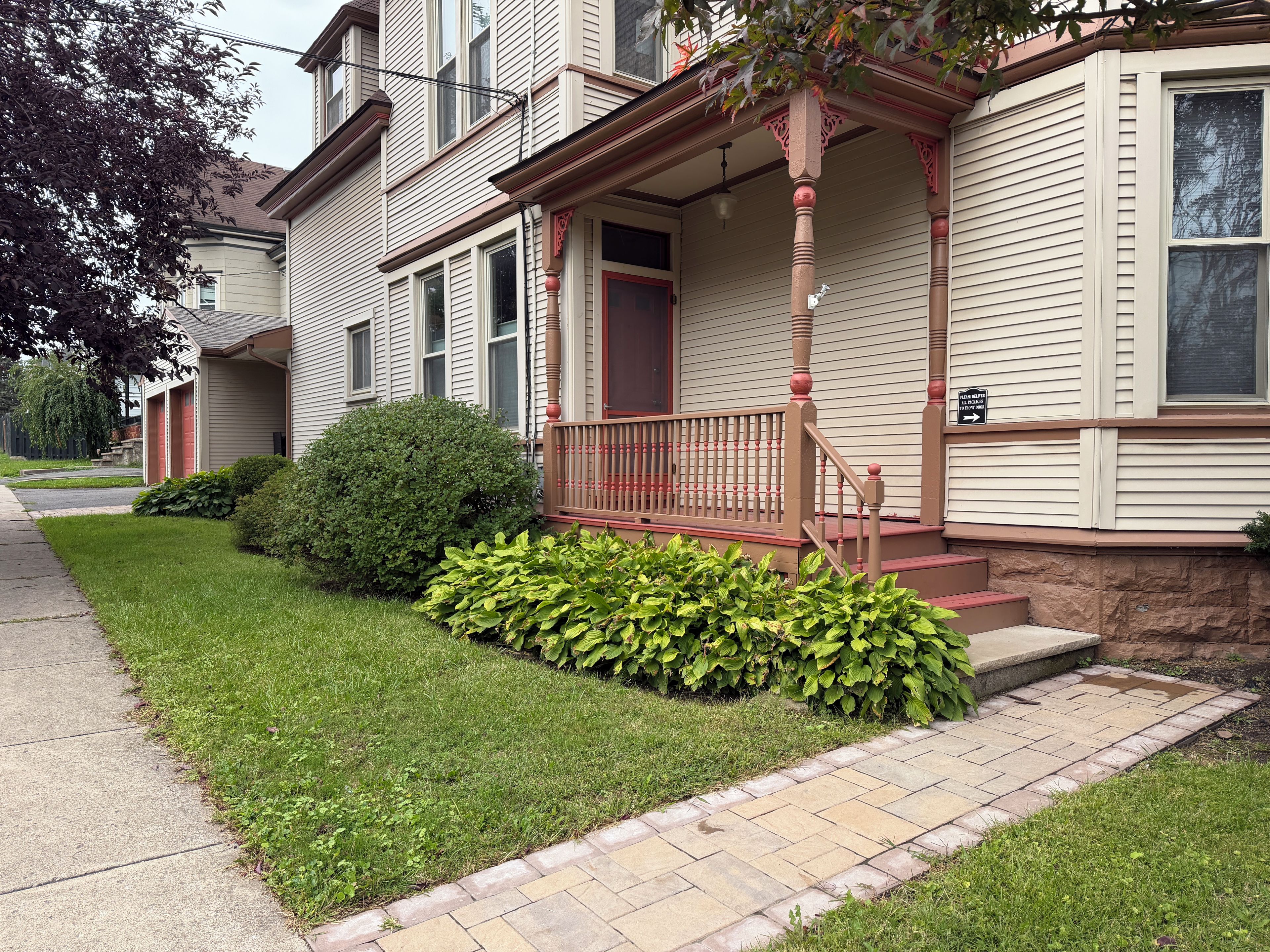A two-story house with a front porch, a small front yard with a hedge, and a brick pathway leading to the door.