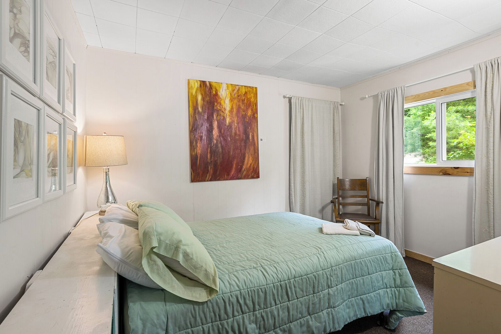 Une chambre à coucher avec un lit, une chaise, un bureau, un tableau et une fenêtre avec des rideaux.