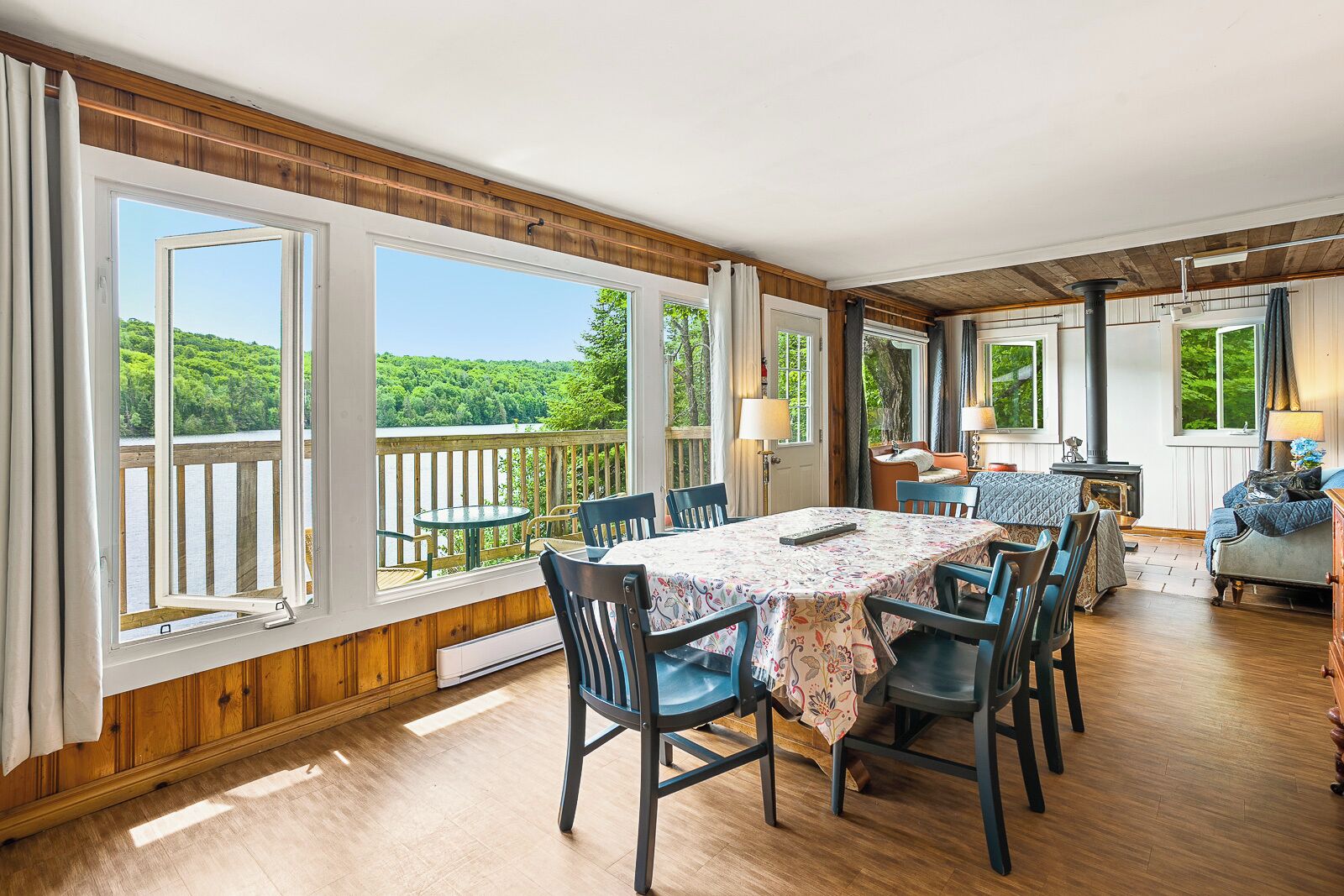 Une pièce chaleureuse avec un plancher en bois, une table à manger avec des chaises, un canapé et un lit. De grandes fenêtres offrent une vue sur une zone boisée.