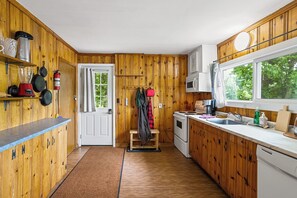 A compact kitchen with wooden walls, a white door, and a window with a view of greenery. - Two Peaceful Lakefront Cottages on beautiful Cardiff Lake. Perfect for families. (Harcourt)