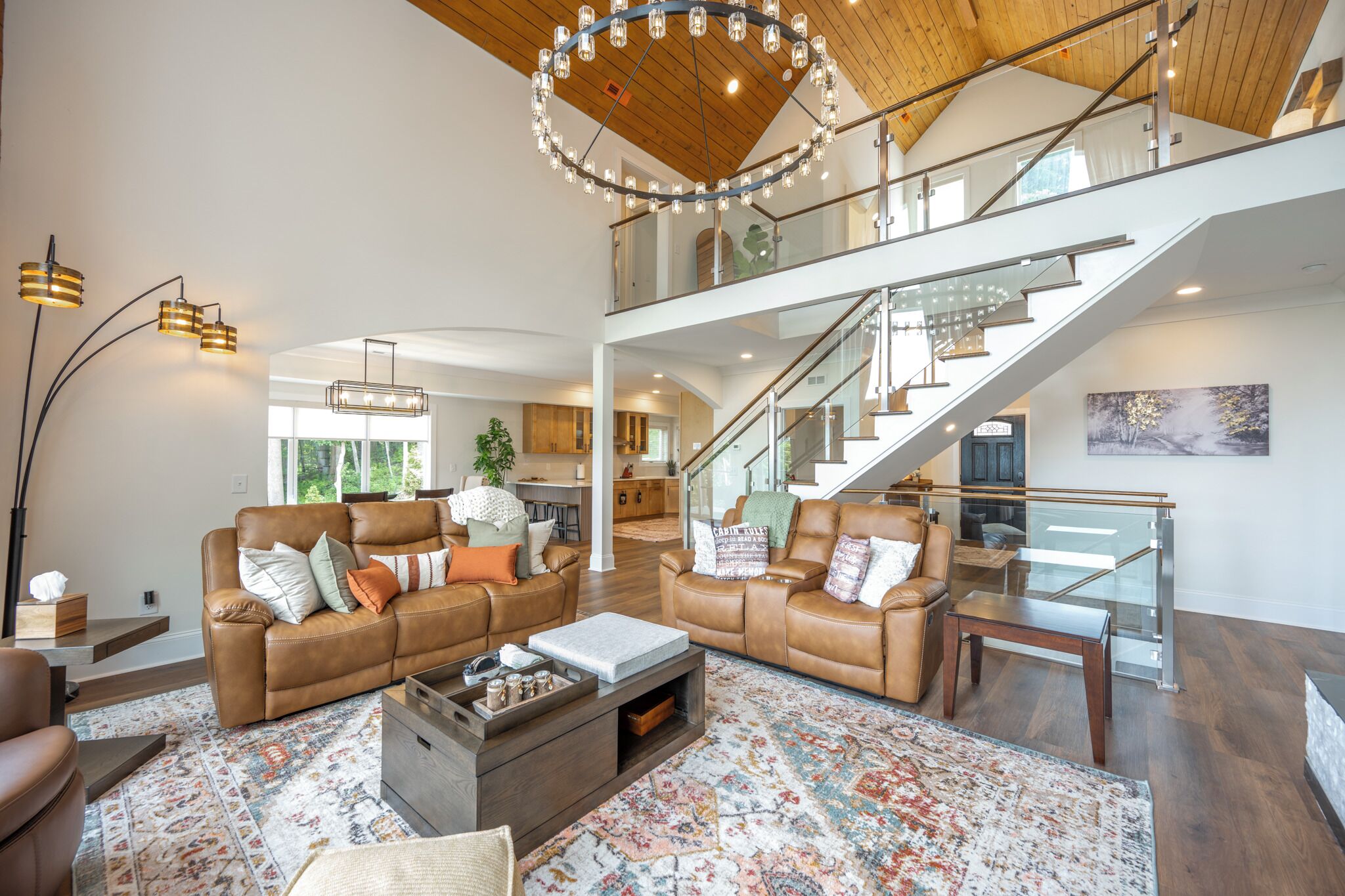 A spacious living area with leather sofas, a coffee table, and a large rug. A staircase with glass railings leads to an upper level.