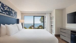 Ein Hotelzimmer mit einem großen Bett, einem Fernseher, einem Balkon mit Meerblick und einem Schreibtisch mit Schubladen.