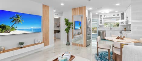 A modern living room with a large flat-screen TV, a dining area with a round table, and a kitchen with white cabinets.