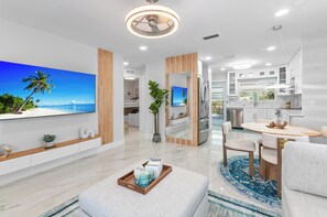 Una sala de estar moderna con un televisor de pantalla plana grande, una zona de comedor con una mesa redonda y una cocina con armarios blancos.