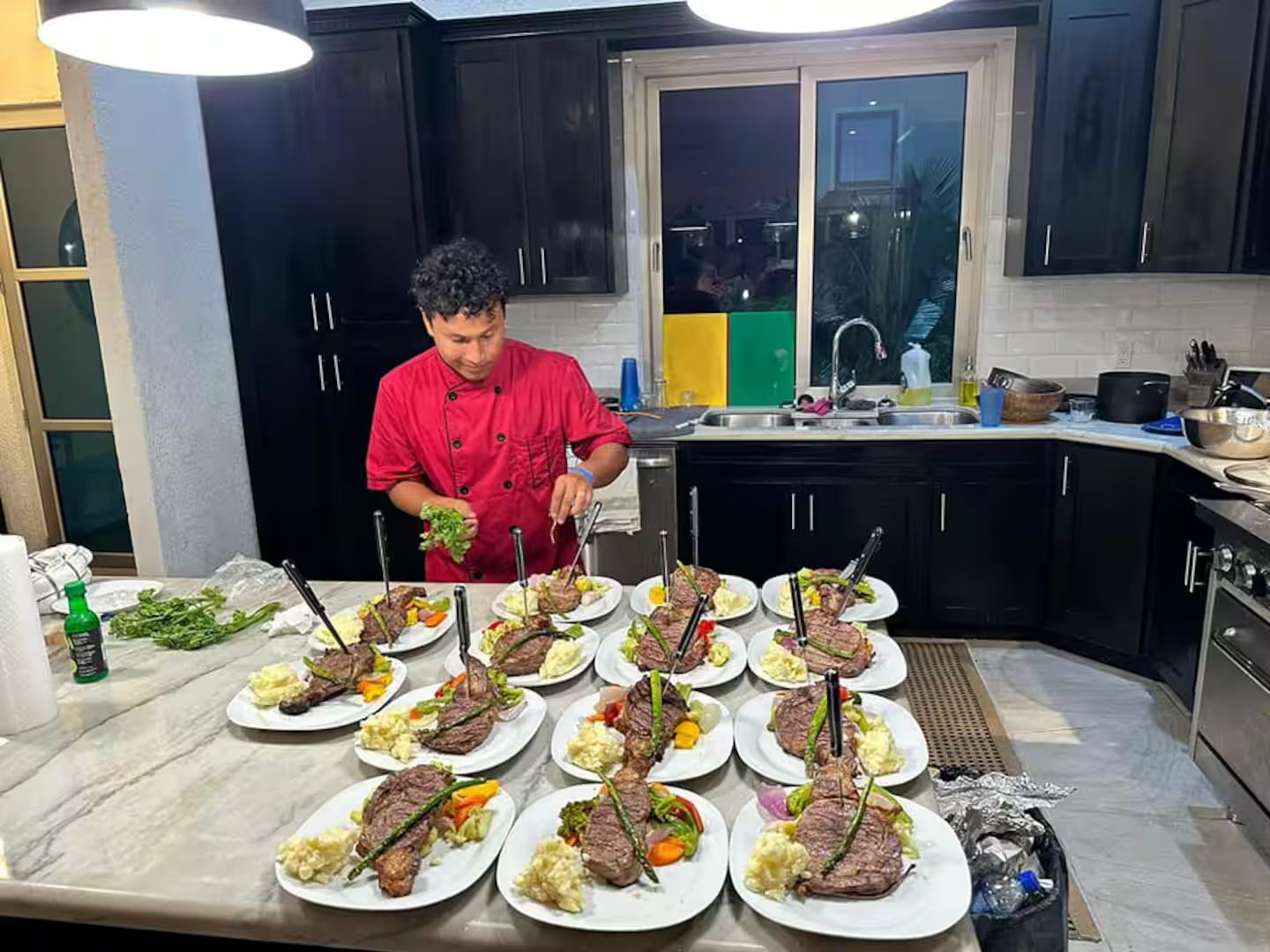 A chef preparing multiple plates of food in a kitchen with black cabinets and a window.