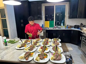 Un chef prepara varios platos de comida en una cocina con gabinetes negros y una ventana.