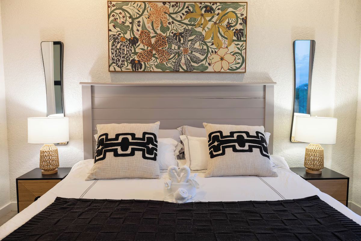 Un lit bien fait, agrémenté de coussins décoratifs, un miroir fixé au mur et, au-dessus, une œuvre d’art aux motifs floraux.